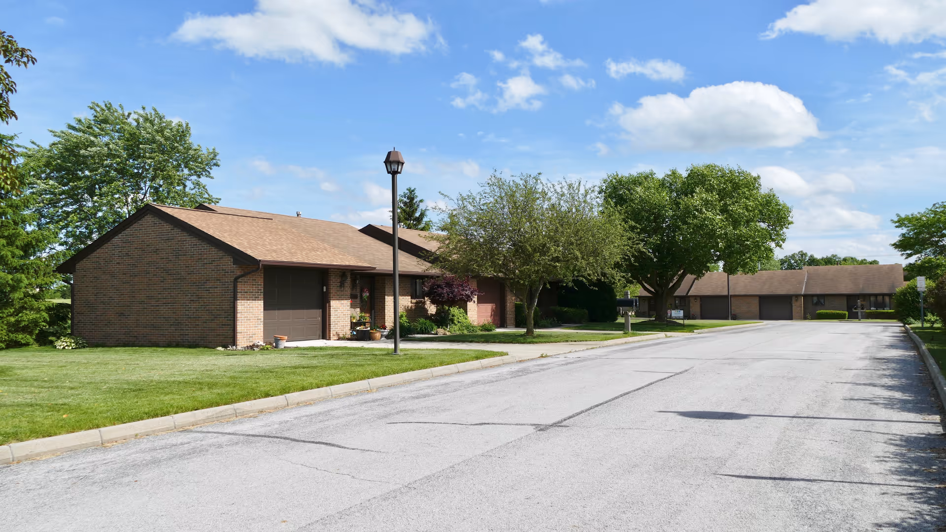 A quiet residential street in a senior living community with single-story brick homes, well-maintained lawns, trees, and a clear blue sky with some clouds.