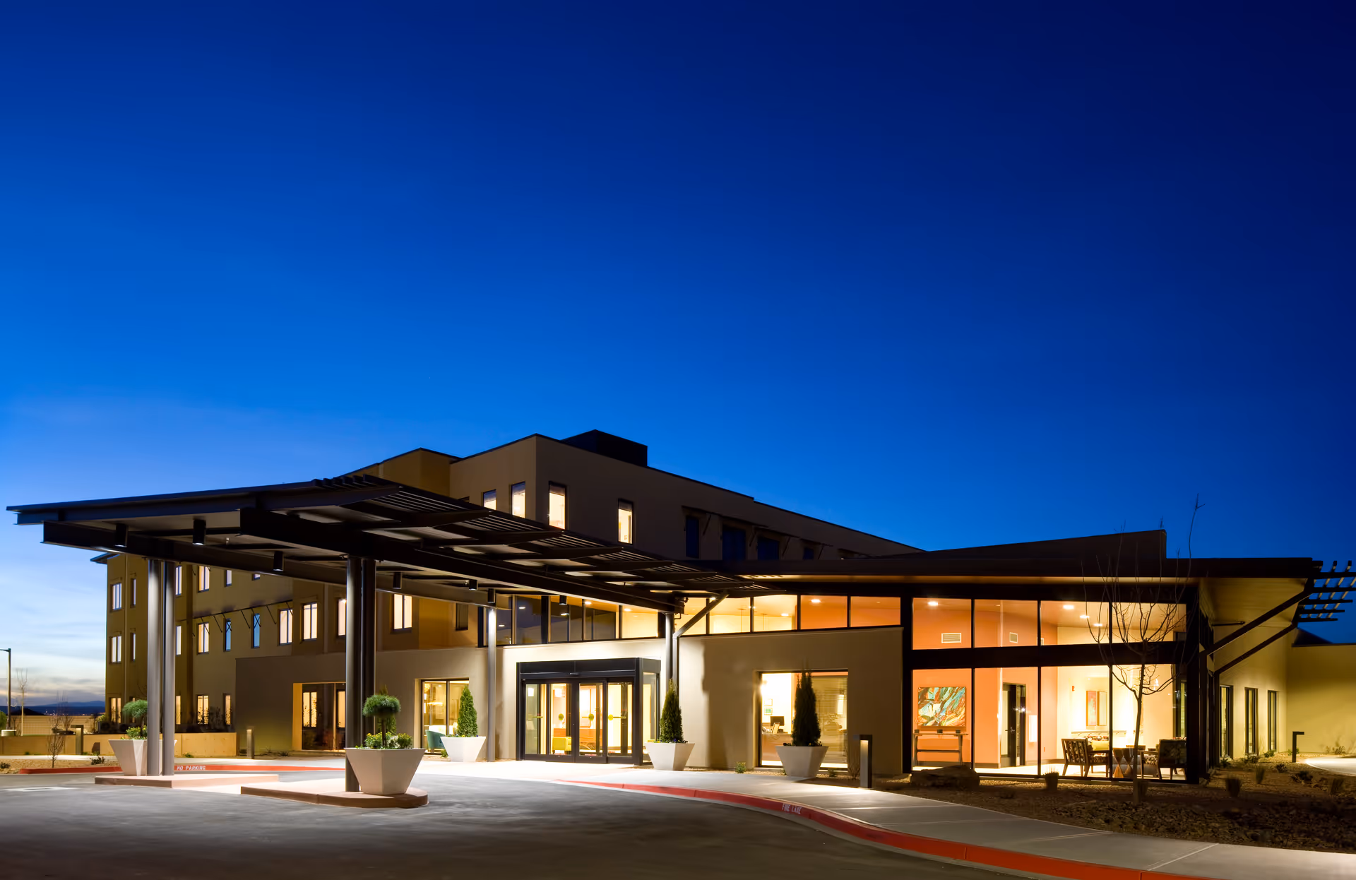 Front exterior of a modern senior living facility at dusk with an illuminated entrance canopy, large windows, and planters.