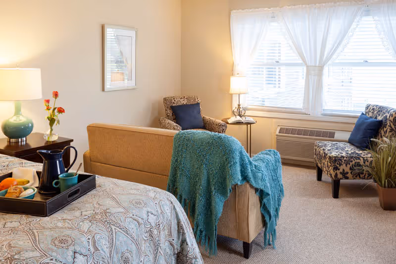 Cozy assisted-living bedroom suite with a bed, sofa, armchairs, and a window with curtains.