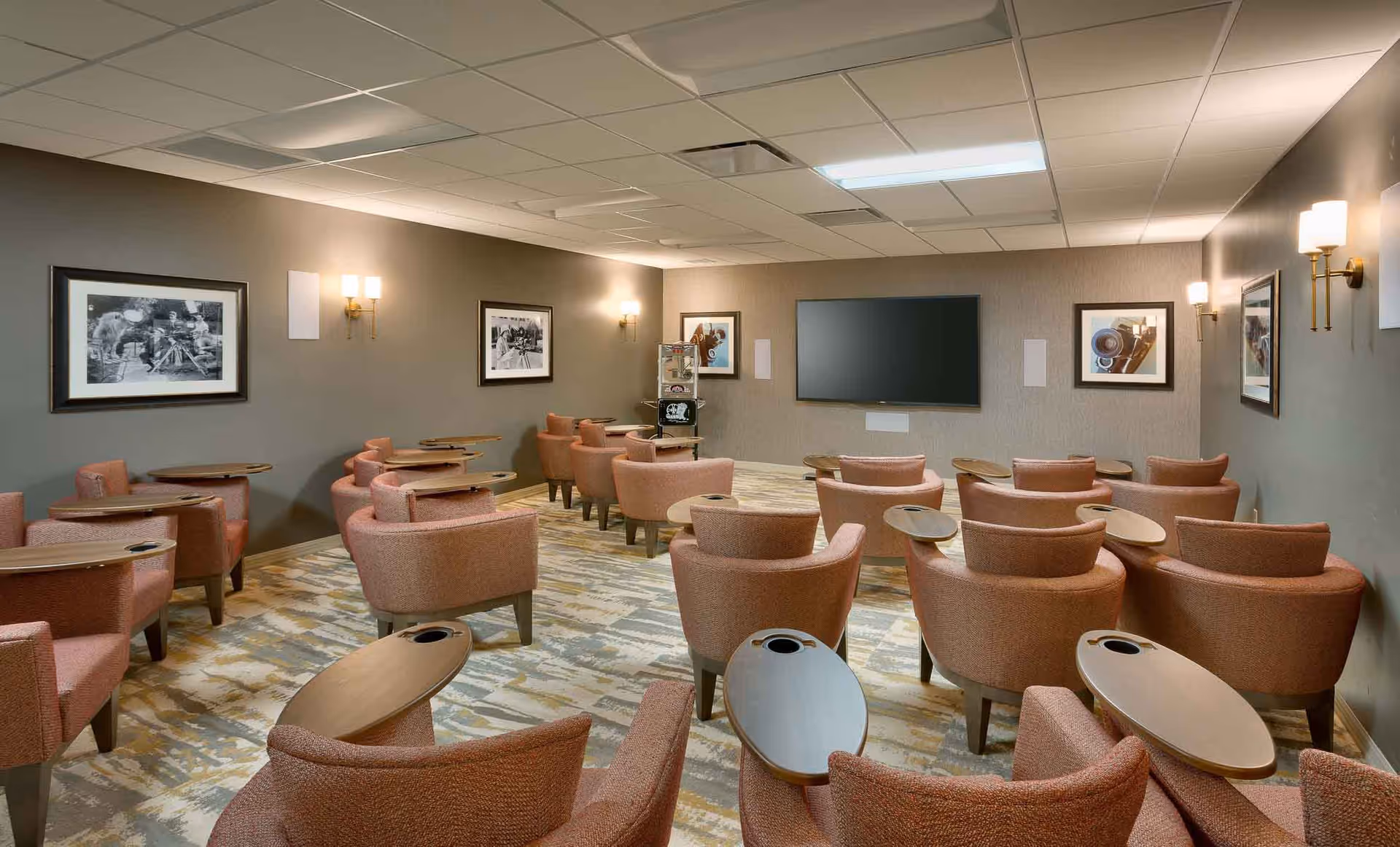 A cozy media room with multiple rows of comfortable armchairs, each equipped with a small attached table. The room has a large flat-screen TV mounted on the front wall, soft lighting from wall sconces, and framed black-and-white photographs on the walls. The carpet has a patterned design in neutral tones.
