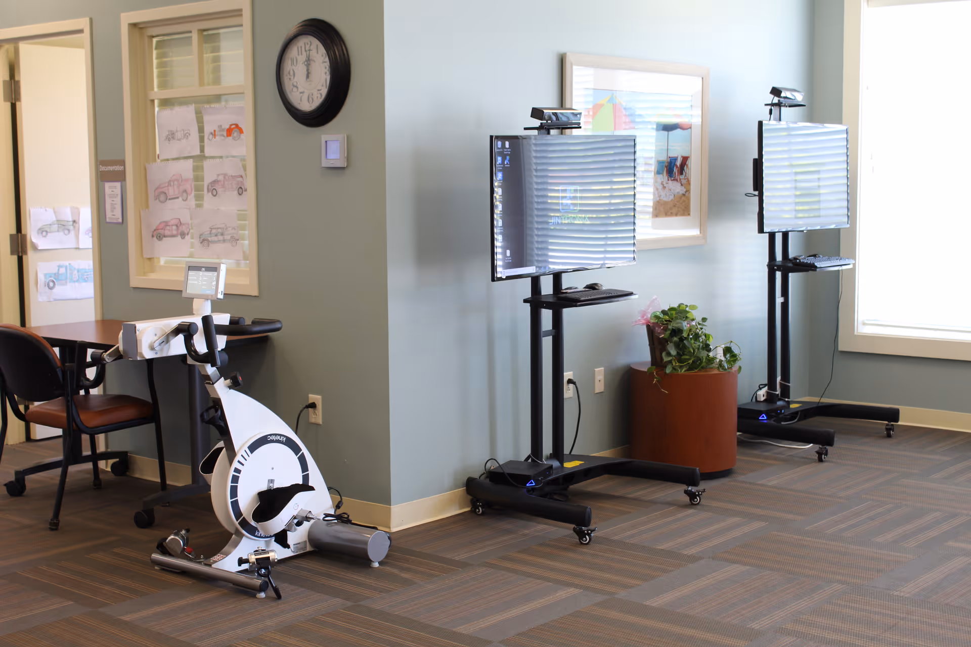 A senior facility common area with an exercise bike, two mobile TV monitors on stands, a potted plant and a table with chairs against a pale green wall.