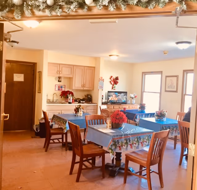 A cozy dining area decorated for the holidays with tables covered in festive tablecloths featuring snowmen. Each table has a small holiday centerpiece with red poinsettias. Wooden chairs surround the tables. In the background, there is a small kitchenette with cabinets, a sink, and holiday decorations. A television is on a stand against the wall, and two windows let in natural light. The ceiling has simple light fixtures, and there is garland with ornaments hanging above the entrance.