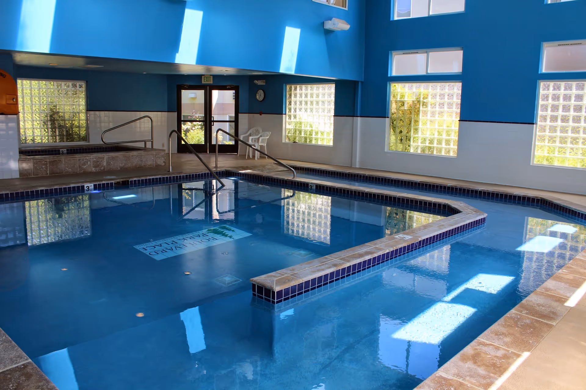 Indoor swimming pool area with blue walls and large windows allowing natural light to enter. The pool has a tiled edge and a handrail leading into the water. There are white chairs near the exit door and a clock on the wall.