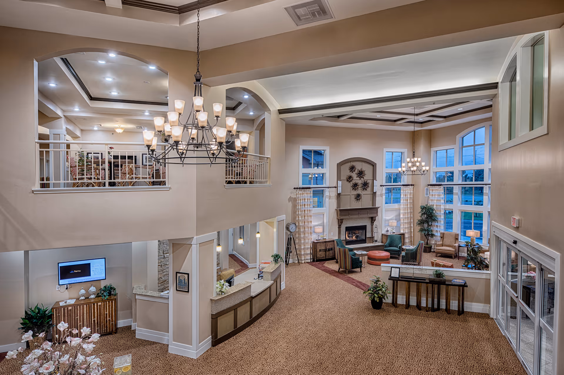 Spacious and well-lit assisted living facility common area with high ceilings, large windows, a fireplace, comfortable seating including armchairs and ottomans, a chandelier, and a reception desk. There is an upper level with additional seating visible through open arches.