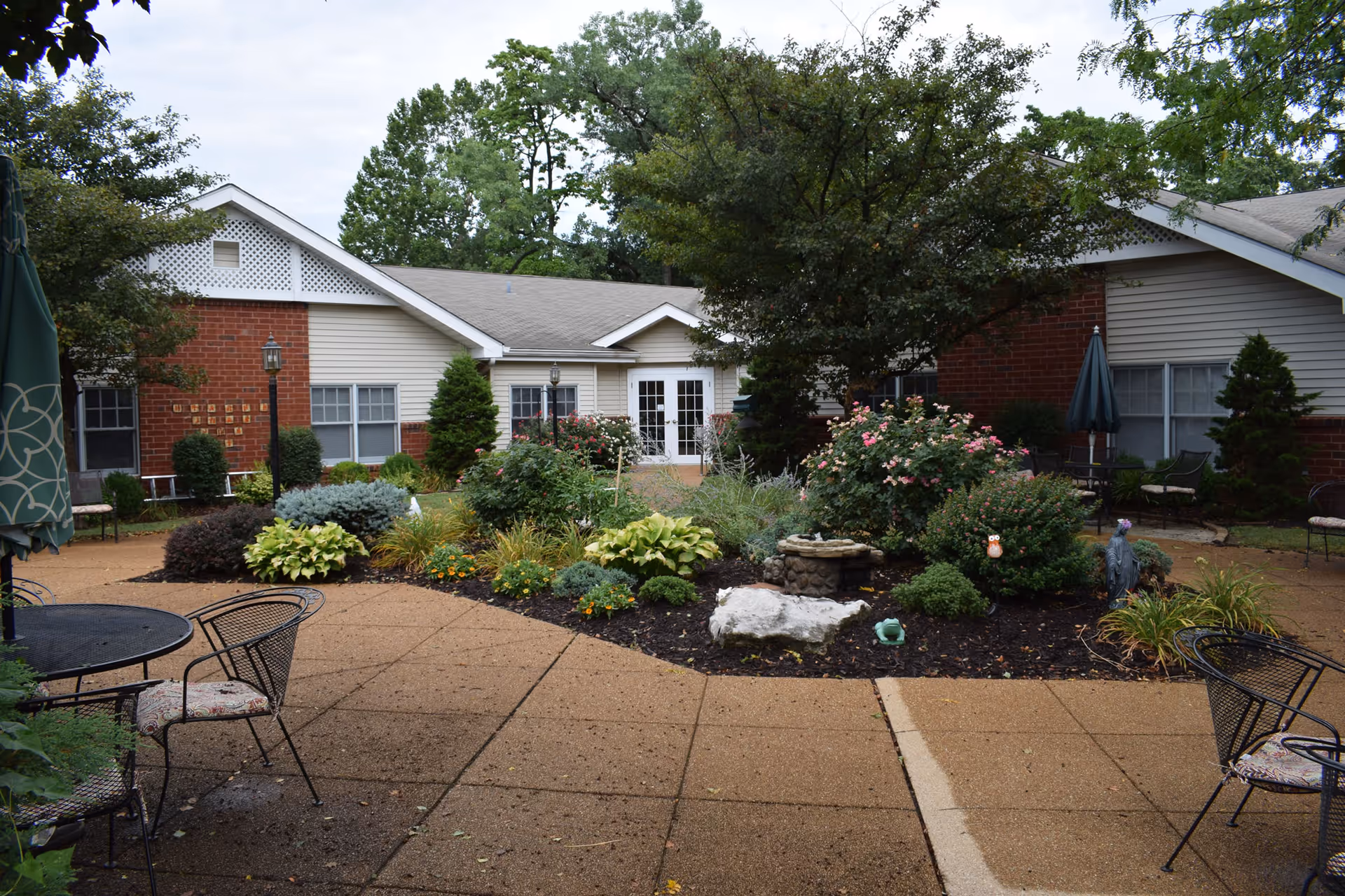 Courtyard garden with patio tables, flower beds, and a fountain in front of a low brick-and-siding residential building.