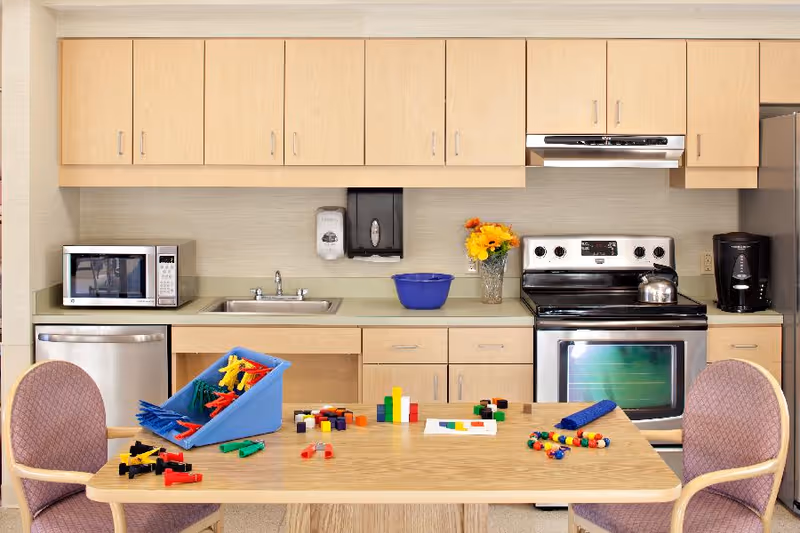 A kitchen area with light wood cabinets, a stainless steel microwave, dishwasher, stove, and refrigerator. On the countertop, there is a blue bowl, a vase with yellow and orange flowers, a coffee maker, and a soap dispenser. In front of the kitchen, there is a wooden table with colorful building blocks and toys scattered on it, flanked by two padded chairs.