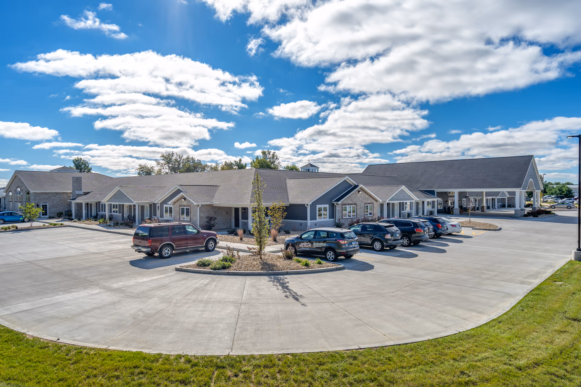Exterior view of Villas of Holly Brook Assisted Living & Memory Care facility in Bethalto, IL, showing a large parking lot with several parked cars, a landscaped island with small trees and shrubs, and a single-story building under a partly cloudy blue sky.