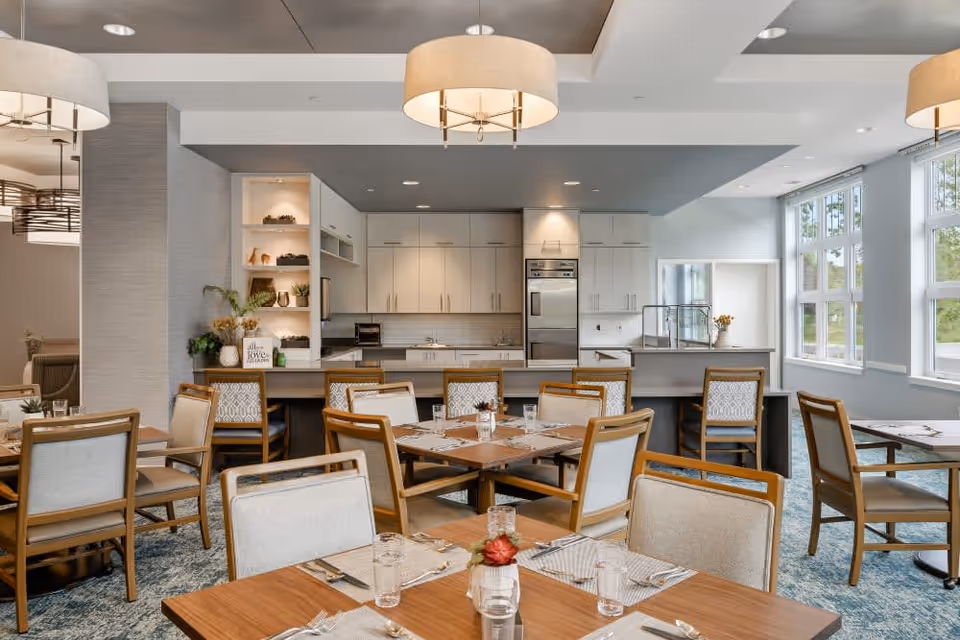 A bright and modern dining area with wooden tables and cushioned chairs set with placemats, glasses, and silverware. The room features large windows letting in natural light, a kitchen area with white cabinets, stainless steel appliances, and decorative shelving. Pendant lights hang from the ceiling, creating a warm and inviting atmosphere.