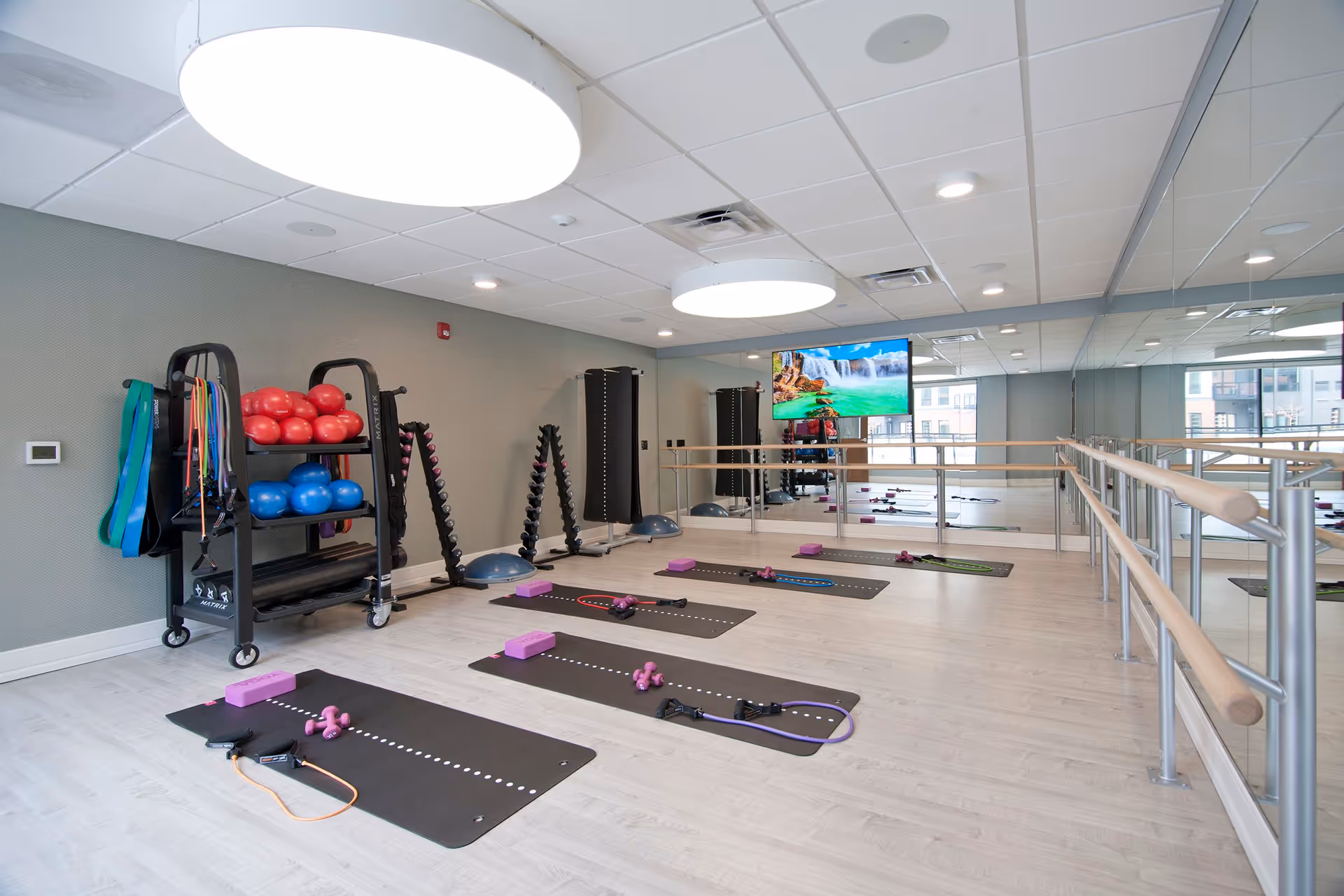 A bright fitness room with light wood flooring and large round ceiling lights. The room has a wall of mirrors and ballet barre rails. On the floor are four black exercise mats, each equipped with purple yoga blocks, small dumbbells, and resistance bands. Against the wall is a rack holding red and blue exercise balls, resistance bands, and other fitness equipment. A flat-screen TV mounted on the mirrored wall displays a nature scene with a waterfall.