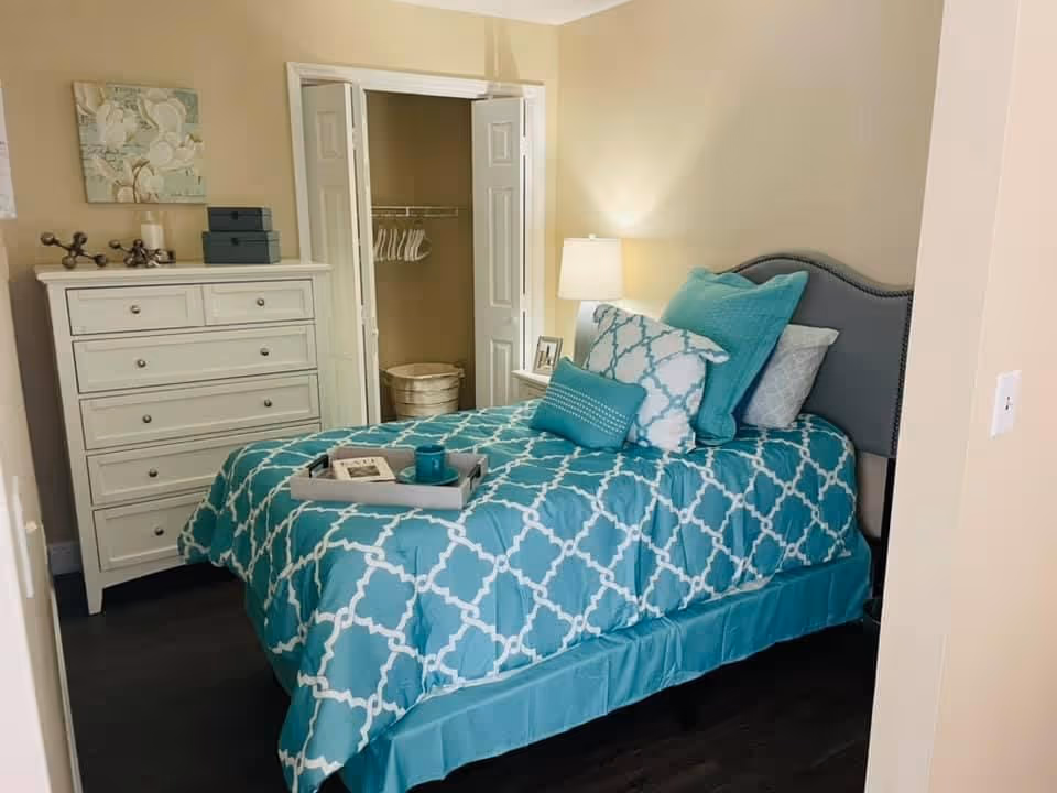 A decorated bedroom with a teal patterned bedspread, upholstered headboard, white dresser, open closet and bedside lamp.