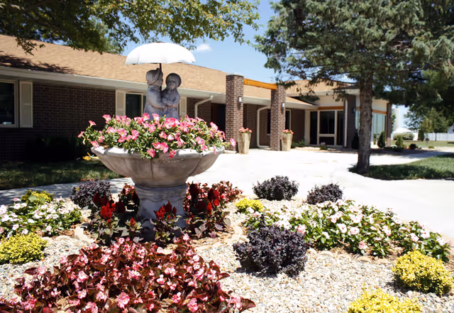 Front entrance of St. Francis Manor showing a flower-filled fountain with a child statue holding an umbrella amid landscaped beds in front of a brick building.