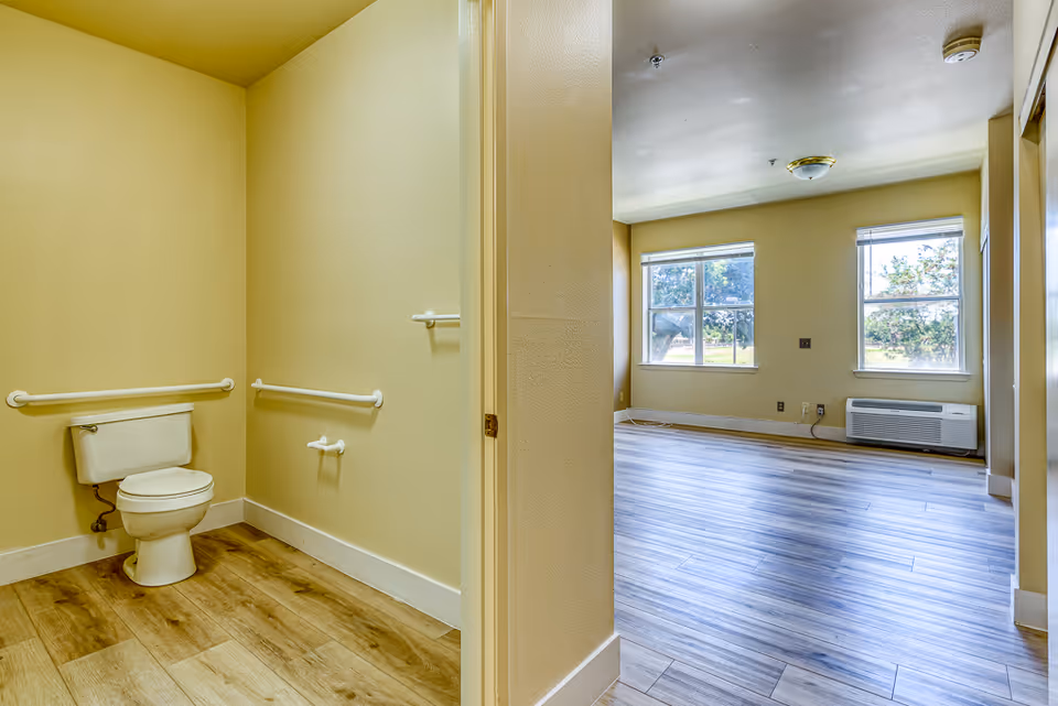 Empty senior living unit interior showing an accessible bathroom with grab bars and an adjacent bright living area with two windows.