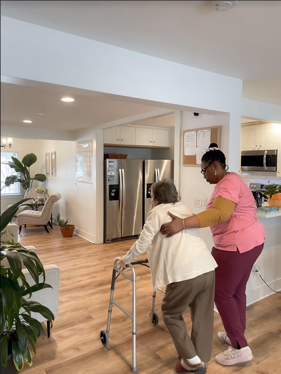 A caregiver in pink scrubs assists an elderly woman using a walker inside a bright, modern living space with wooden floors, plants, and a kitchen area with stainless steel appliances in the background.