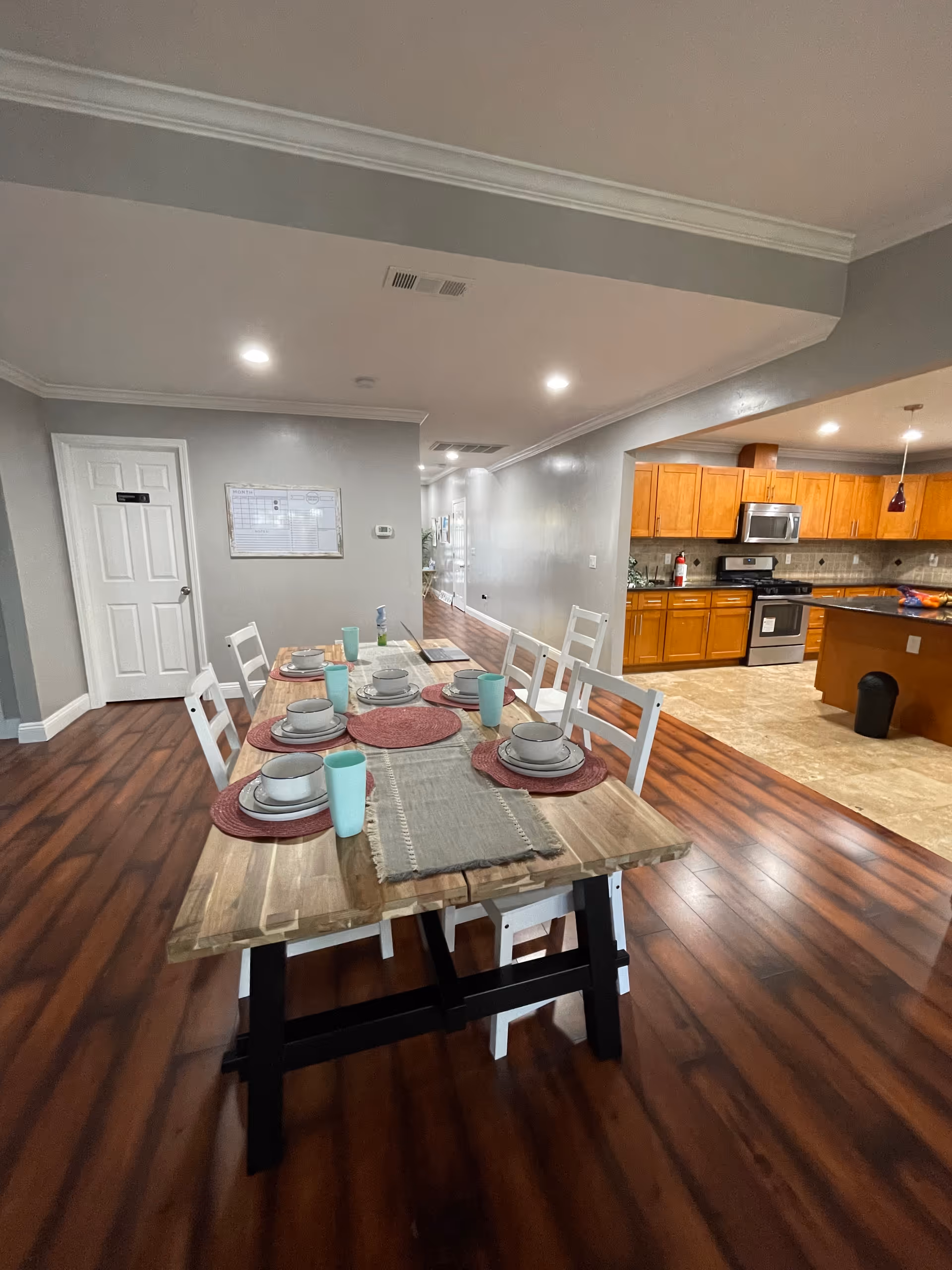 Interior view of a senior living facility dining area with a wooden table set for six people with plates, bowls, and cups. The room has wooden flooring and gray walls with white trim. To the right, there is an open kitchen with wooden cabinets, stainless steel appliances, and a tiled floor. The ceiling has recessed lighting.