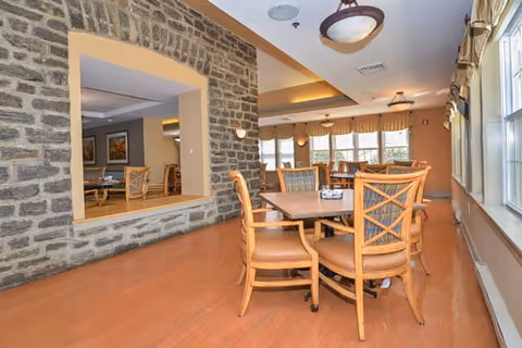 A dining area with wooden tables and chairs featuring cushioned seats. The room has large windows with curtains allowing natural light to fill the space. A stone wall with a large rectangular opening separates this area from another room with additional seating and artwork on the walls. Ceiling lights provide additional illumination.