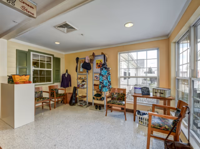 A bright room with large windows letting in natural light, featuring wooden chairs with floral cushions arranged around the space. There is a small table with a basket and a sign, a white pedestal with a basket on top, and a display rack holding various hats, bags, and clothing items. The walls are painted yellow and white, and there is a window with green shutters on one side.