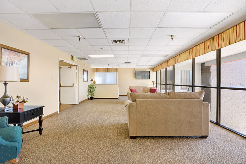 A spacious senior living facility common area with beige carpet and walls, featuring two beige sofas with pink cushions facing a wall-mounted TV. Large windows with yellow valances let in natural light. A green armchair and a black side table with a lamp and decorative items are on the left side. A framed painting and a potted plant are also visible.