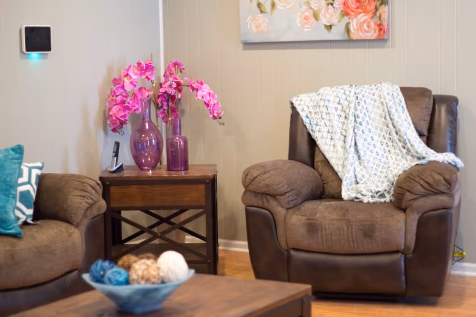 A cozy living room area with a brown cushioned armchair draped with a light blue patterned throw blanket. Next to it is a wooden side table holding two purple vases with pink flowers and a cordless phone. Part of a brown sofa with blue and white patterned pillows is visible on the left. A wooden coffee table with decorative balls in a blue bowl is in the foreground. The wall behind is beige with a floral painting hanging above the armchair.