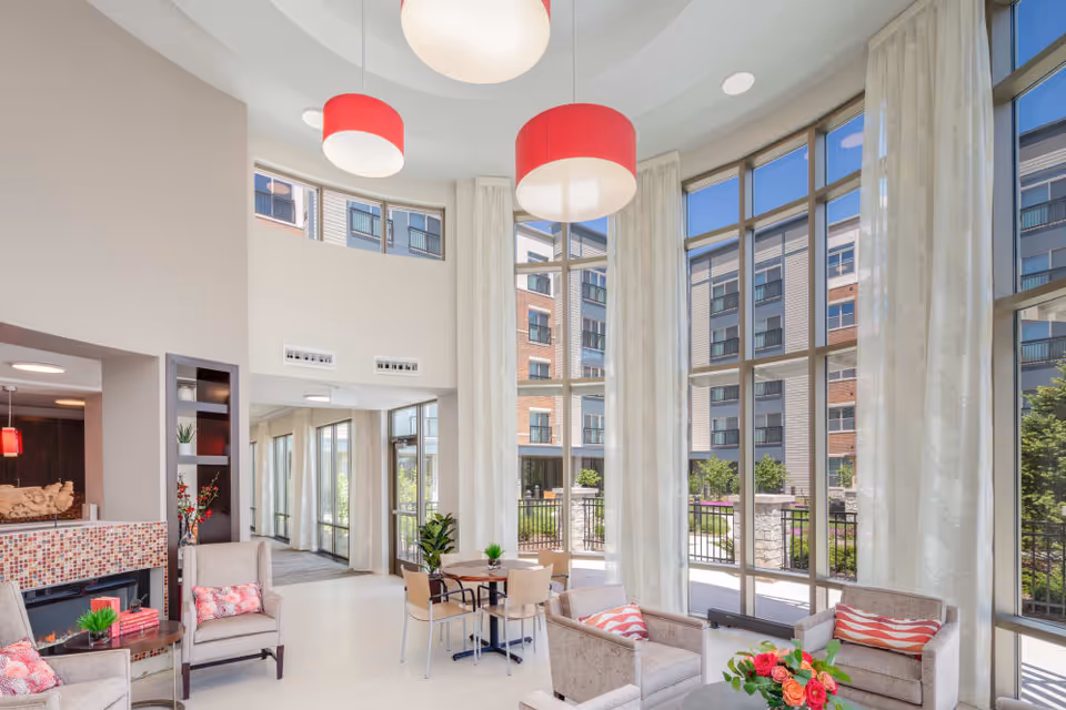Bright and spacious living room area with large floor-to-ceiling windows allowing natural light to fill the space. The room features comfortable armchairs with patterned cushions, a small round table with chairs, a fireplace with mosaic tile surround, and modern pendant lights hanging from the ceiling. Outside the windows, a courtyard with greenery and a multi-story building are visible.
