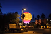 A nighttime outdoor scene at Westminster Village featuring a brightly lit hot air balloon in yellow, pink, and purple colors. The area is surrounded by palm trees, buildings, and street lamps illuminating the pathway.