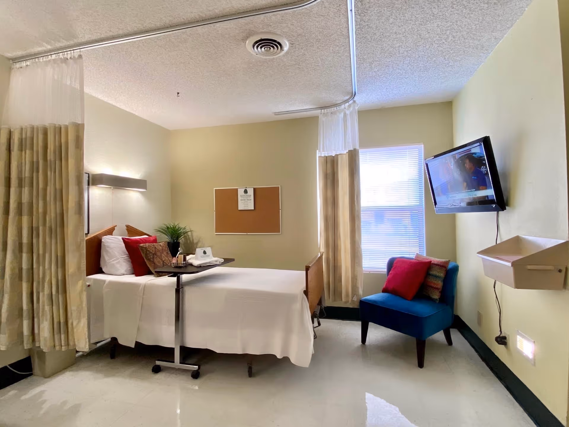 A nursing facility room with a hospital bed covered in white sheets and a tray table with a plant and some items on it. There is a blue armchair with red and patterned pillows near a window with blinds. A flat-screen TV is mounted on the wall opposite the bed. Beige privacy curtains hang from a ceiling track around the bed.