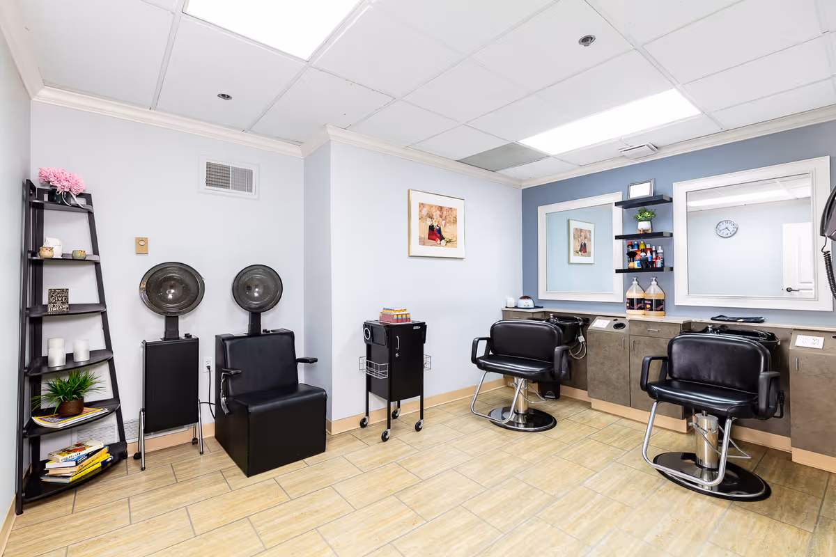 A clean and well-lit salon room with two black salon chairs in front of mirrors and a washing station. There are two hair dryer chairs against the wall, a black shelving unit with decorative items and books, and a small rolling cart with supplies. The walls are painted light blue and white, and the floor is tiled.