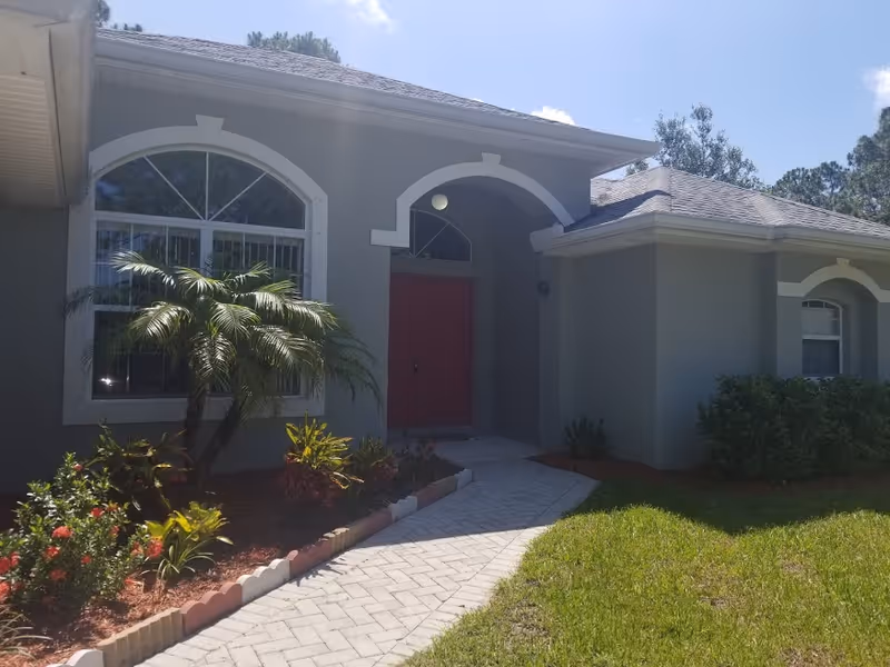 Front exterior of a single-story grey stucco house with a red front door, arched windows, a paved walkway, and landscaped plants including a small palm.