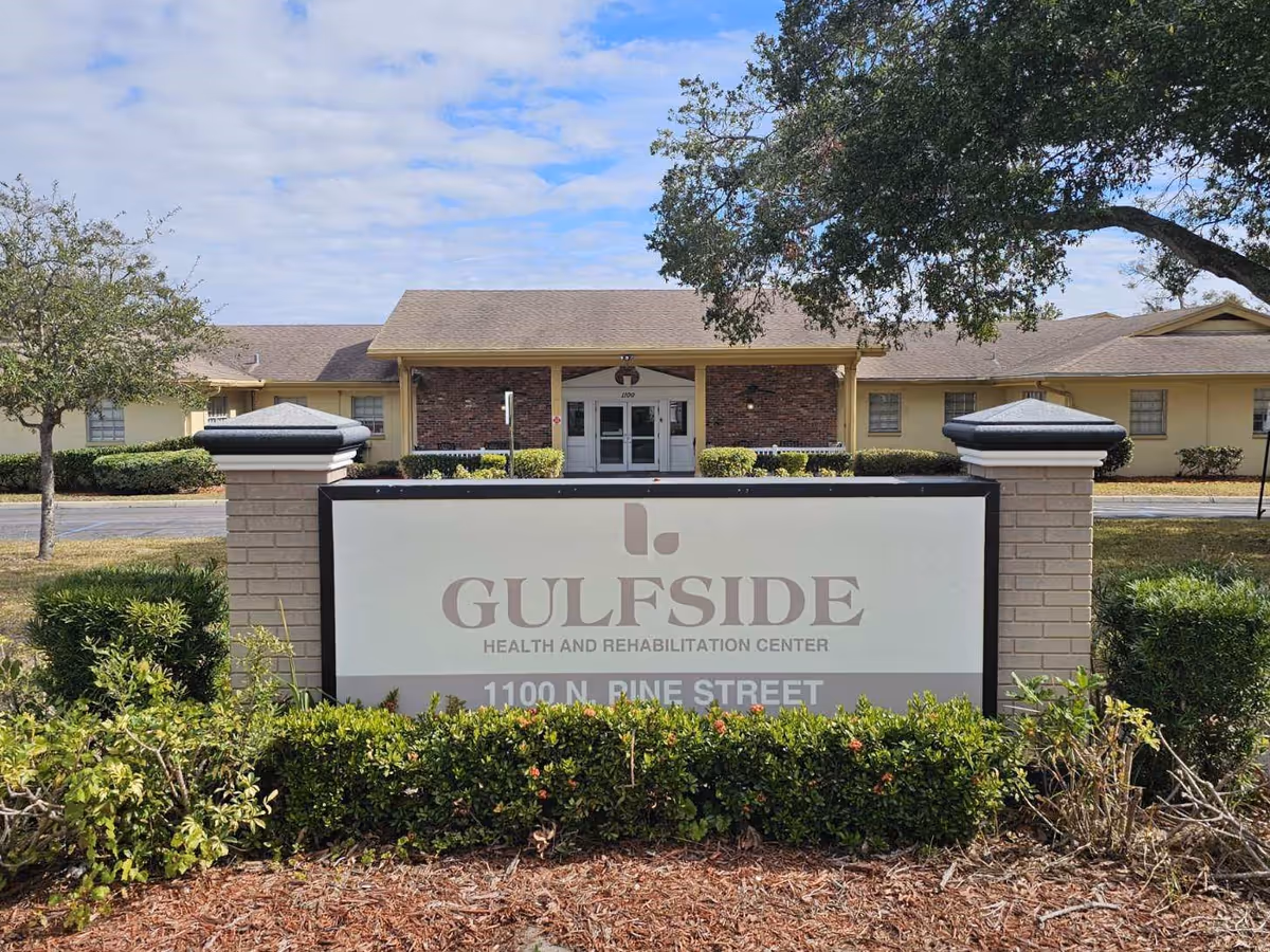 Exterior view of Gulfside Health and Rehabilitation Center showing the main entrance of the building behind a large sign with the facility's name and address, surrounded by trees and shrubs under a partly cloudy sky.