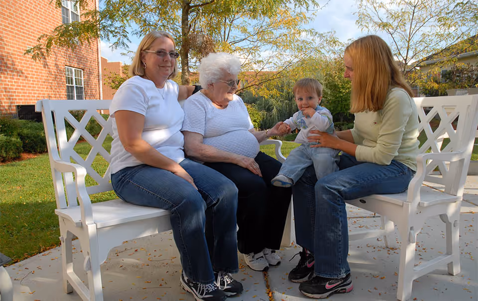 Three women and a toddler sitting on white benches outside in a garden area with trees and a brick building in the background. The elderly woman in the center is holding the toddler's hand while the other two women sit on either side, smiling and interacting with the child.
