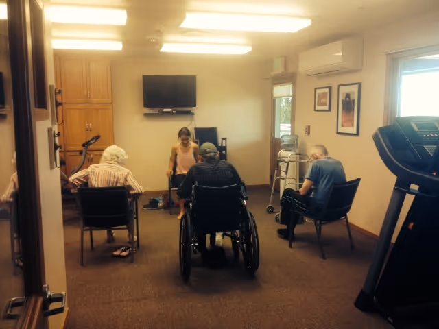 A small group of elderly individuals seated in chairs and a wheelchair in a well-lit room with beige walls and carpeted floor. A young woman is engaging with them, possibly leading an activity. The room contains exercise equipment including a treadmill and stationary bike, a wall-mounted TV, and framed pictures on the walls.