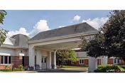 Covered entrance/porte-cochère of a low-rise brick building with landscaped grounds under a partly cloudy sky.