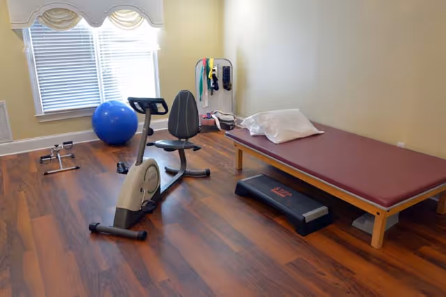 Small exercise/therapy room with a recumbent bike, exercise ball, step platform, treatment table, and resistance bands by a window.