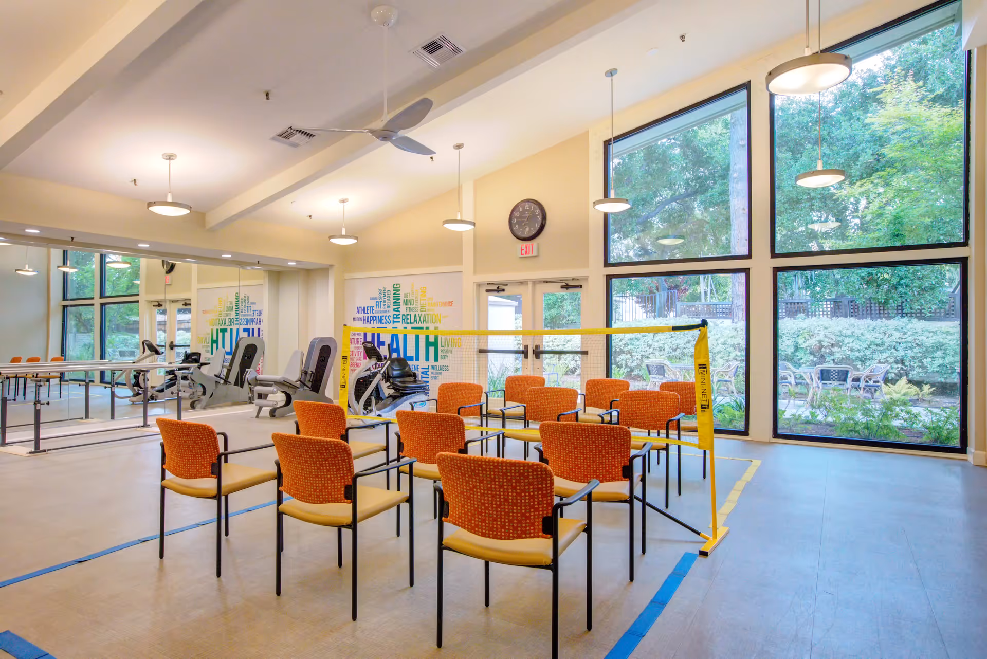 A bright exercise room with large windows showing outdoor greenery. The room contains several orange and yellow chairs arranged in rows behind a yellow caution tape barrier. Exercise equipment, including stationary bikes and parallel bars, is visible near a mirrored wall. The walls have colorful word art related to health and wellness.