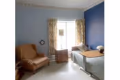 A small, cozy room with light blue walls and a darker blue accent wall. The room contains a brown armchair, a hospital-style bed with a wooden overbed table, a small wooden nightstand with a lamp, and floral curtains covering a window.
