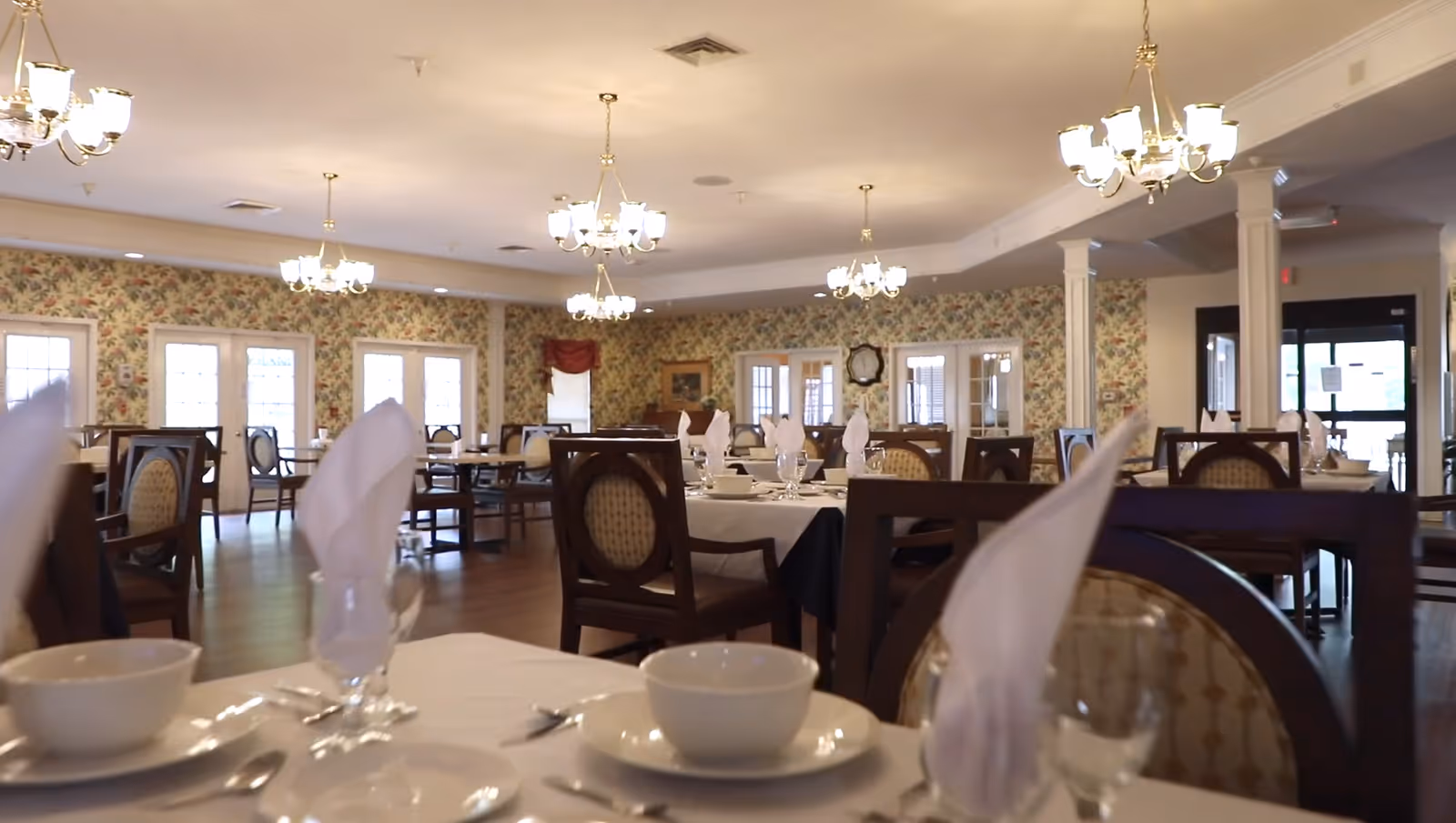 Spacious dining room with set tables, chandeliers, and floral wallpaper.