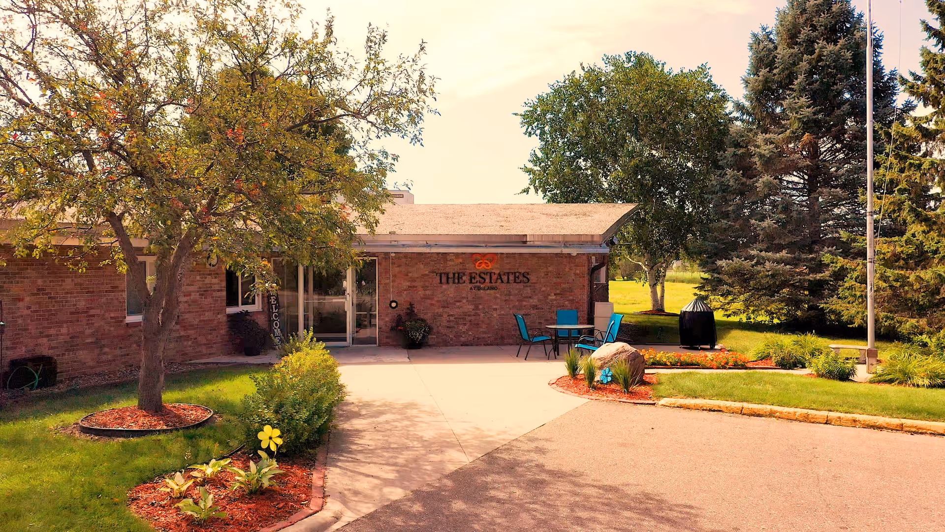 Exterior view of The Estates at Delano facility showing a brick building entrance with a tree and landscaped garden beds with flowers and shrubs. There is a small patio area with blue chairs and a table near the entrance.