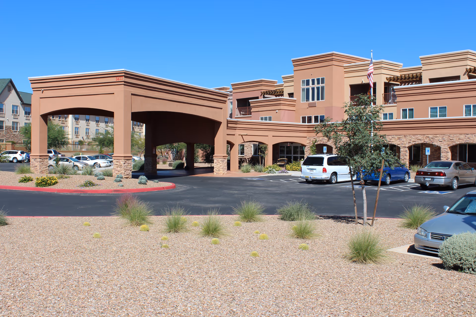 Exterior view of a senior living facility with a covered entrance, parking lot with several cars, desert landscaping with gravel and small bushes, and a clear blue sky.
