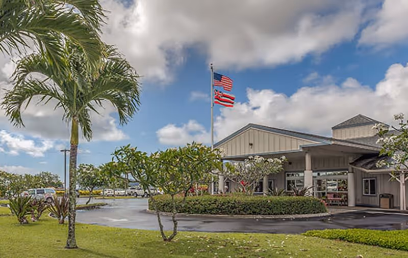 Exterior view of Regency at Puakea facility showing a single-story building with a covered entrance, surrounded by green landscaping including palm trees and bushes, with American and Hawaiian flags flying on a flagpole against a partly cloudy sky.
