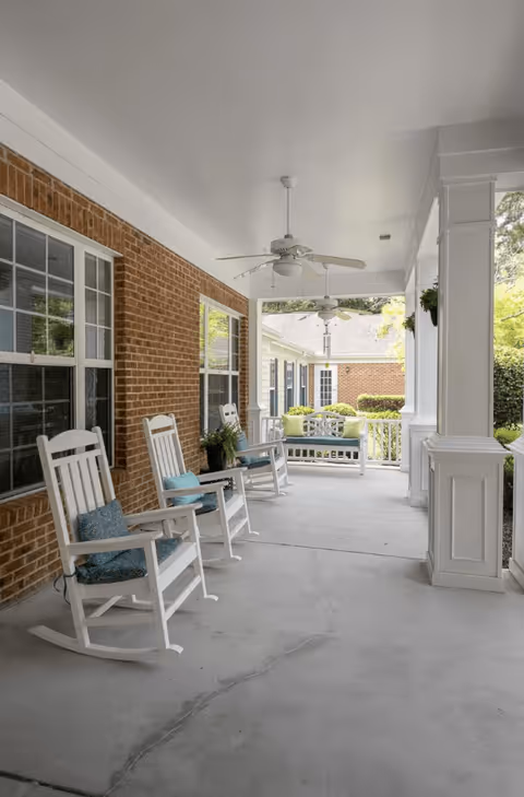 Covered outdoor porch area with white rocking chairs and a bench with cushions, ceiling fans, brick wall on one side, and white pillars supporting the roof. Greenery and other buildings are visible in the background.