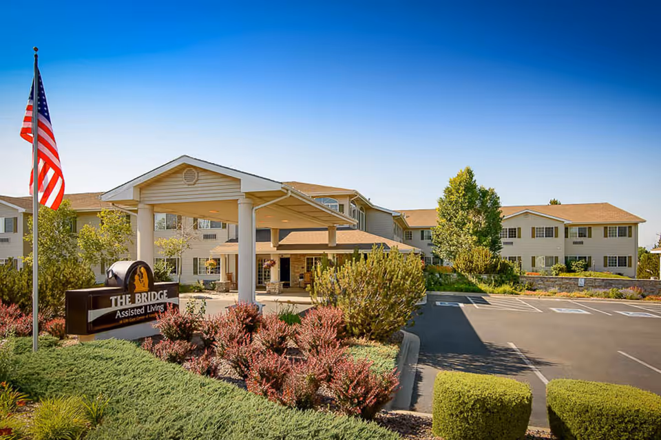 Front entrance of The Bridge at Longmont assisted living facility with a covered porte-cochère, landscaped beds, an American flag, and a parking lot.