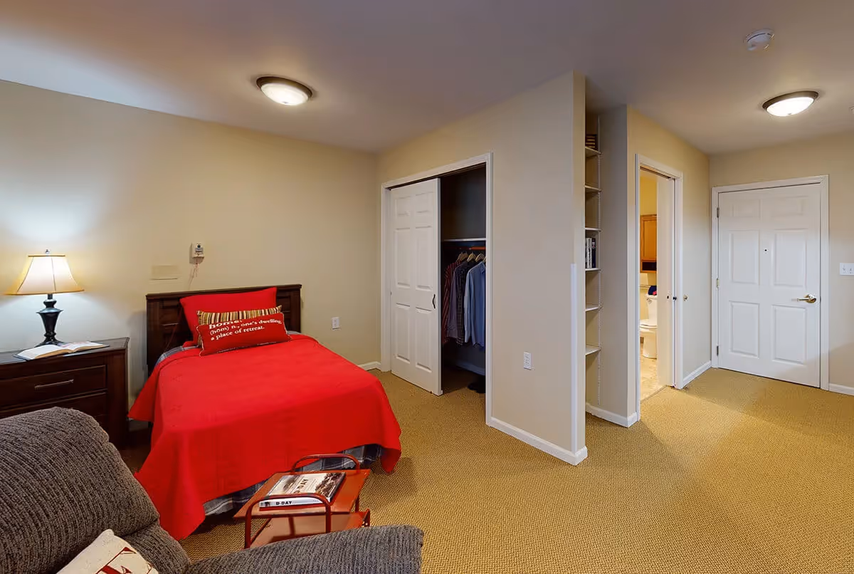 A furnished bedroom with a red bedspread, nightstand and lamp, open closet, and doors leading to a bathroom and hallway.