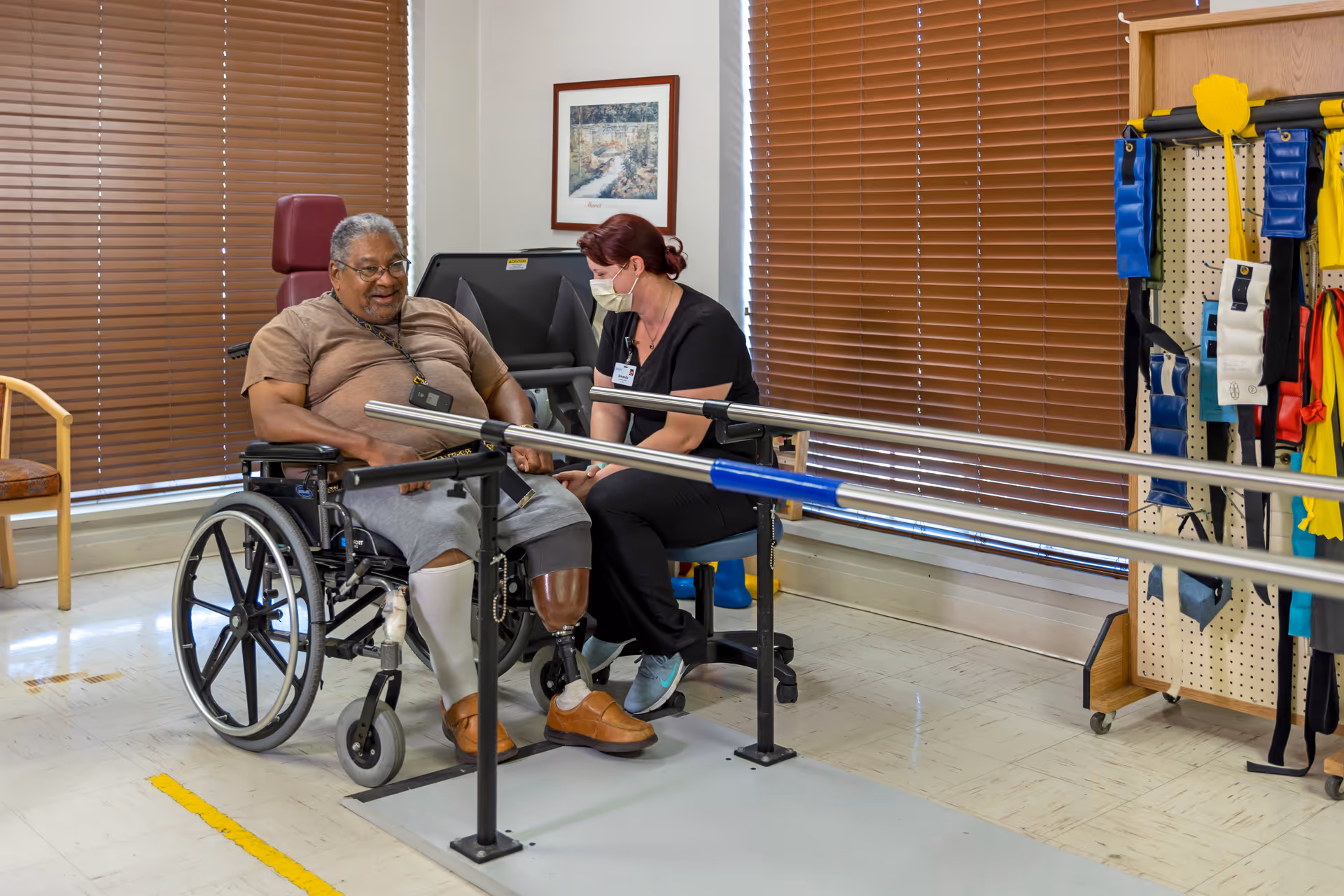A man in a wheelchair with a prosthetic leg is receiving physical therapy from a female therapist wearing a mask in a rehabilitation room. The room has parallel bars for walking exercises, a chair, brown window blinds, and various therapy equipment hanging on a pegboard.
