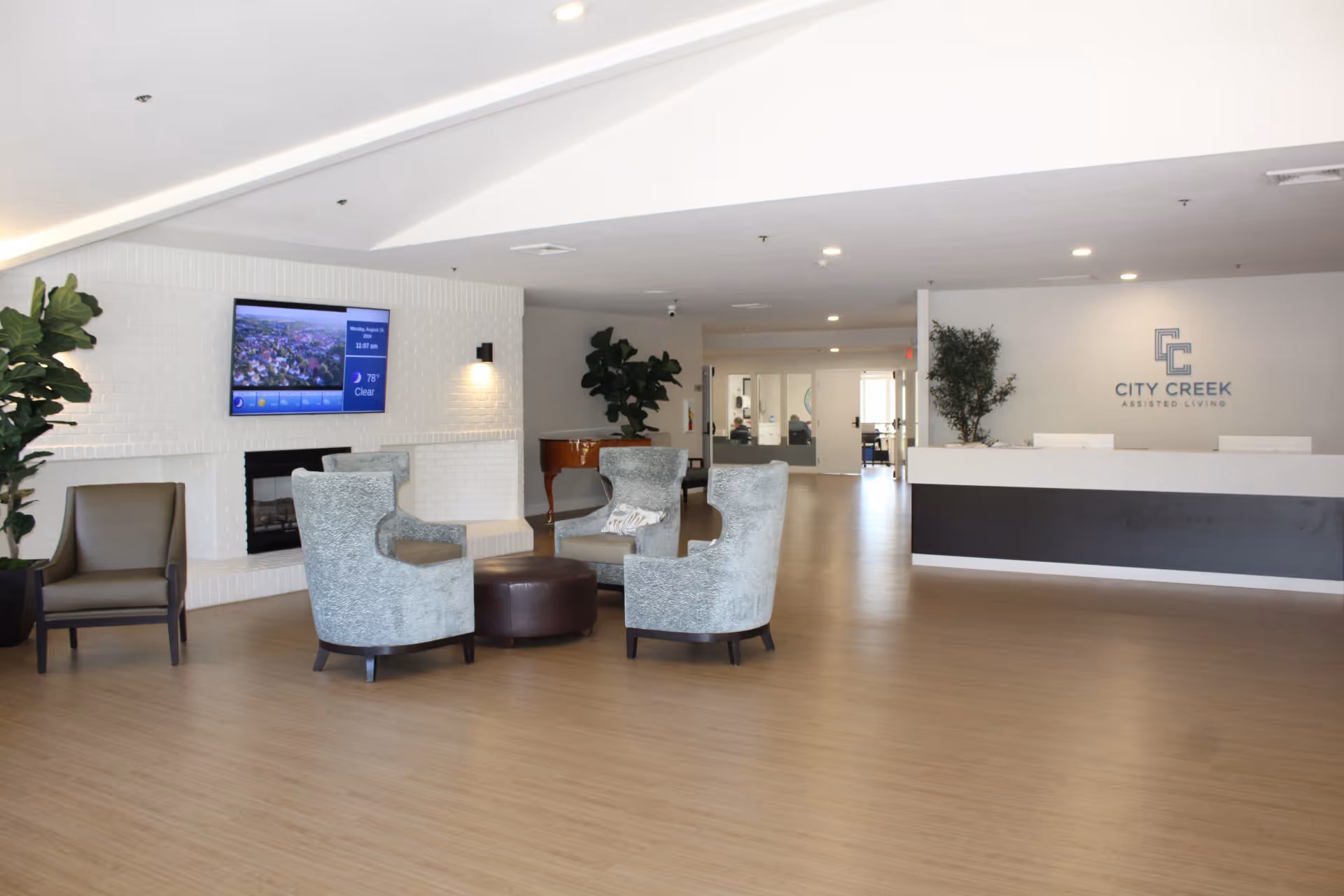 A spacious and modern assisted living facility lobby with a seating area consisting of four armchairs around a round ottoman, a white brick fireplace with a mounted flat-screen TV above it, potted plants, and a reception desk with the City Creek Assisted Living logo on the wall behind it.