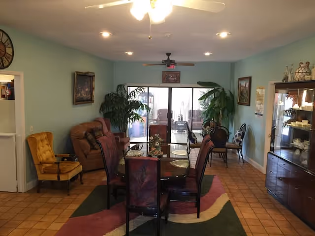 A cozy living room area with a round dining table surrounded by six chairs on a colorful rug. The room has light blue walls, a ceiling fan with lights, and several potted plants near a large sliding glass door leading to an outdoor patio. There are additional seating options including a yellow armchair and a brown recliner, as well as framed artwork on the walls and a glass-front cabinet displaying dishes.