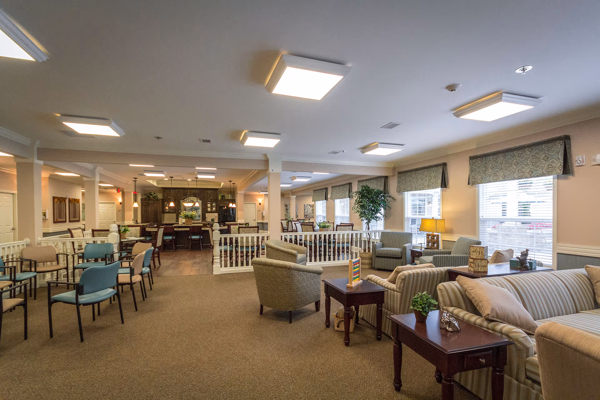 A spacious and well-lit common area in a senior living facility featuring multiple seating arrangements including striped sofas, armchairs, and dining chairs. The room has large windows with patterned valances, several side tables with decorative items and plants, and a kitchen area with dark wood cabinetry and pendant lighting in the background.