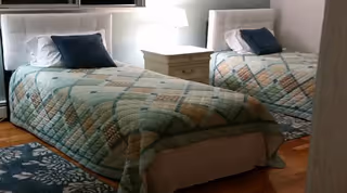 Two twin beds with patterned quilts, upholstered headboards and a shared nightstand with a lamp in a bedroom.