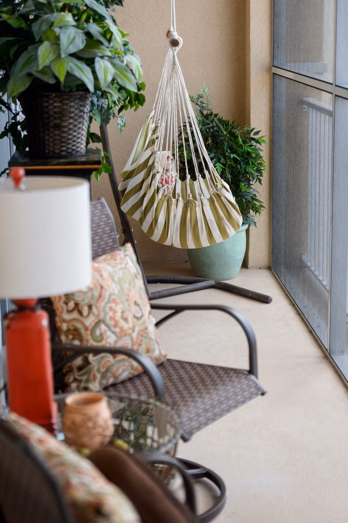 A cozy screened balcony with a hanging striped hammock chair, wicker seating with patterned cushions, potted plants, and a small table with a lamp.