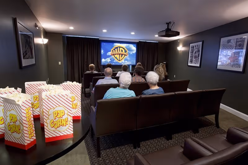 Elderly residents sit in rows of seats watching a movie in a small theater room with popcorn boxes on a table in the foreground.