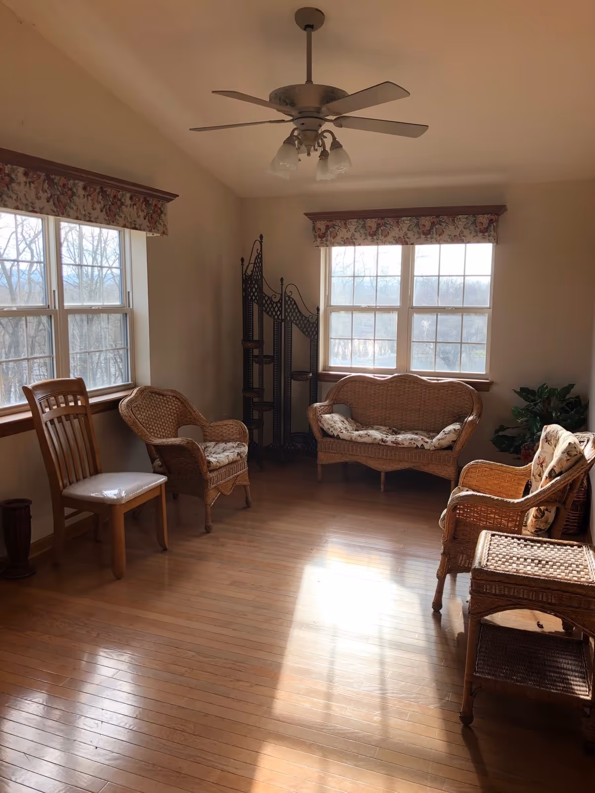 A sunlit room with wooden floors featuring wicker furniture including a loveseat, two armchairs with floral cushions, a wicker side table, and a wooden chair with a white cushion. Two large windows with floral valances allow natural light to fill the space. A ceiling fan with lights hangs from the ceiling, and a decorative black metal folding screen is placed in the corner near a green potted plant.