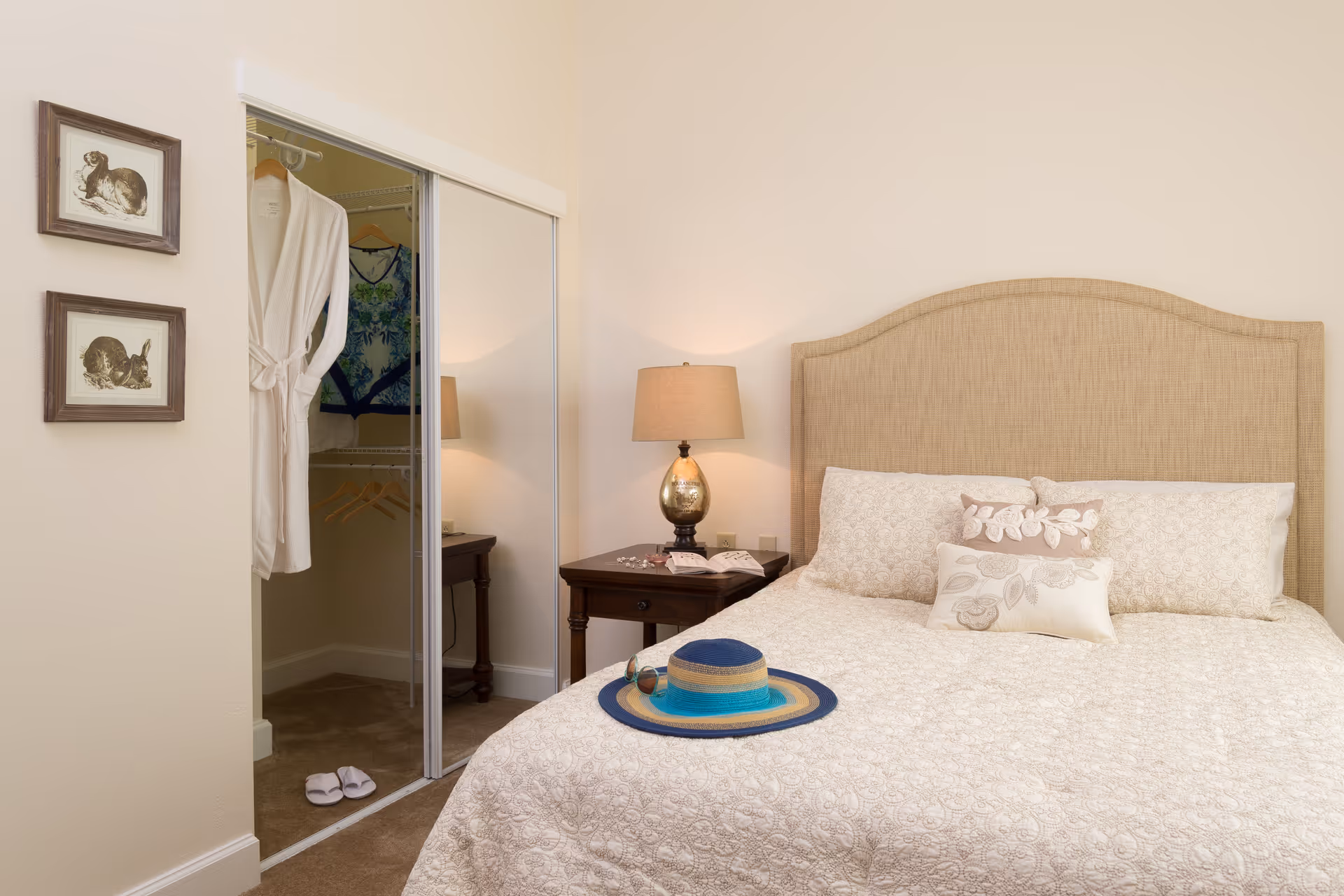 A cozy bedroom with a beige upholstered headboard and a bed covered in a light patterned quilt with decorative pillows. A blue and beige sunhat and sunglasses rest on the bed. To the left, there is a nightstand with a lamp and an open book. A closet with sliding mirrored doors is partially open, revealing a white robe and some hanging clothes. Two framed pictures of rabbits hang on the wall beside the closet.